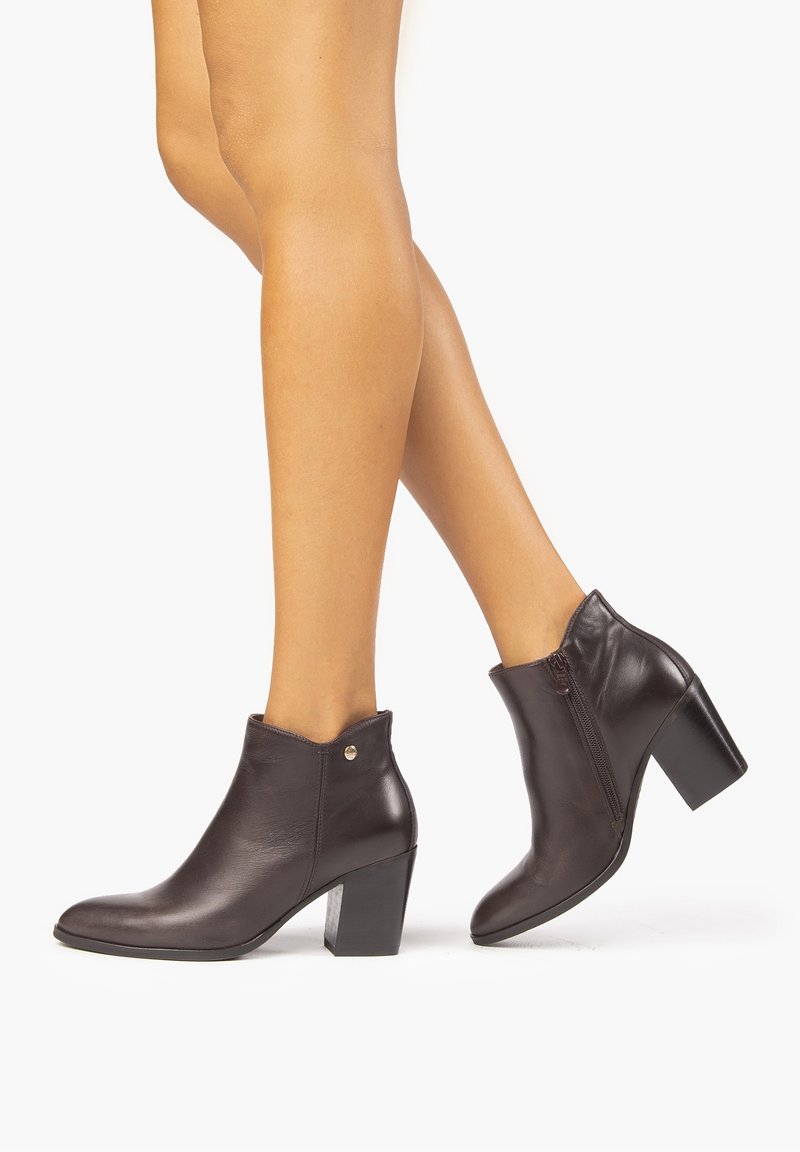Personne portant des bottines en cuir marron foncé à talons épais, debout sur un fond blanc avec les jambes croisées à la cheville.