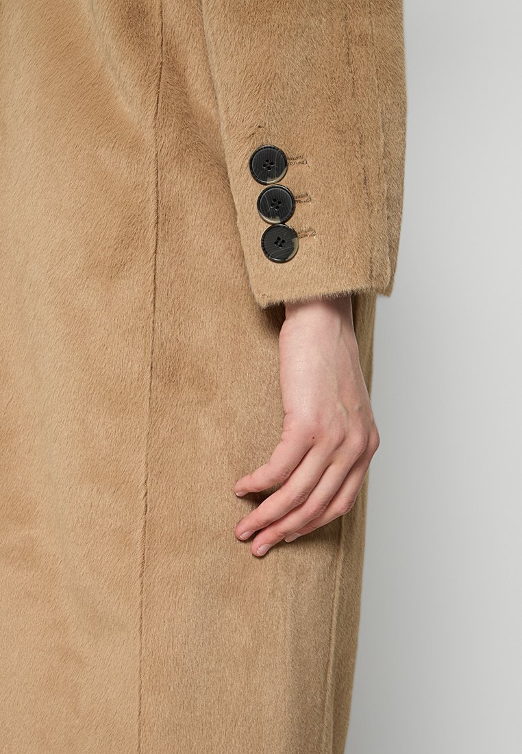 Karmelowy płaszcz z sztucznego futra z trzema czarnymi guzikami na rękawie, o gładkiej teksturze i prostym kroju.