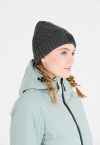 Grijze gebreide beanie met een gemarmerd patroon en een zwart leren logo label, gecombineerd met een lichtblauwe ritsjack met capuchon.