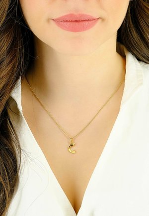 Femme portant un collier en or avec un pendentif en forme de croissant, vêtue d'un haut blanc, montrant les lèvres et le haut de la poitrine.