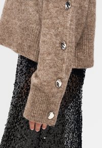 Καφέ πλεκτό πουλόβερ με υφή ριμπ και στρογγυλά ασημένια κουμπιά στο μανίκι, συνδυασμένο με μαύρο ένδυμα με παγιέτες.