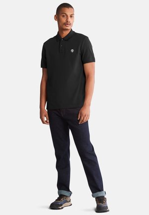 Zwarte polo met een korte kraag, klein logo op de linkerkant van de borst, gedragen met donkerblauwe jeans en grijze wandelschoenen.