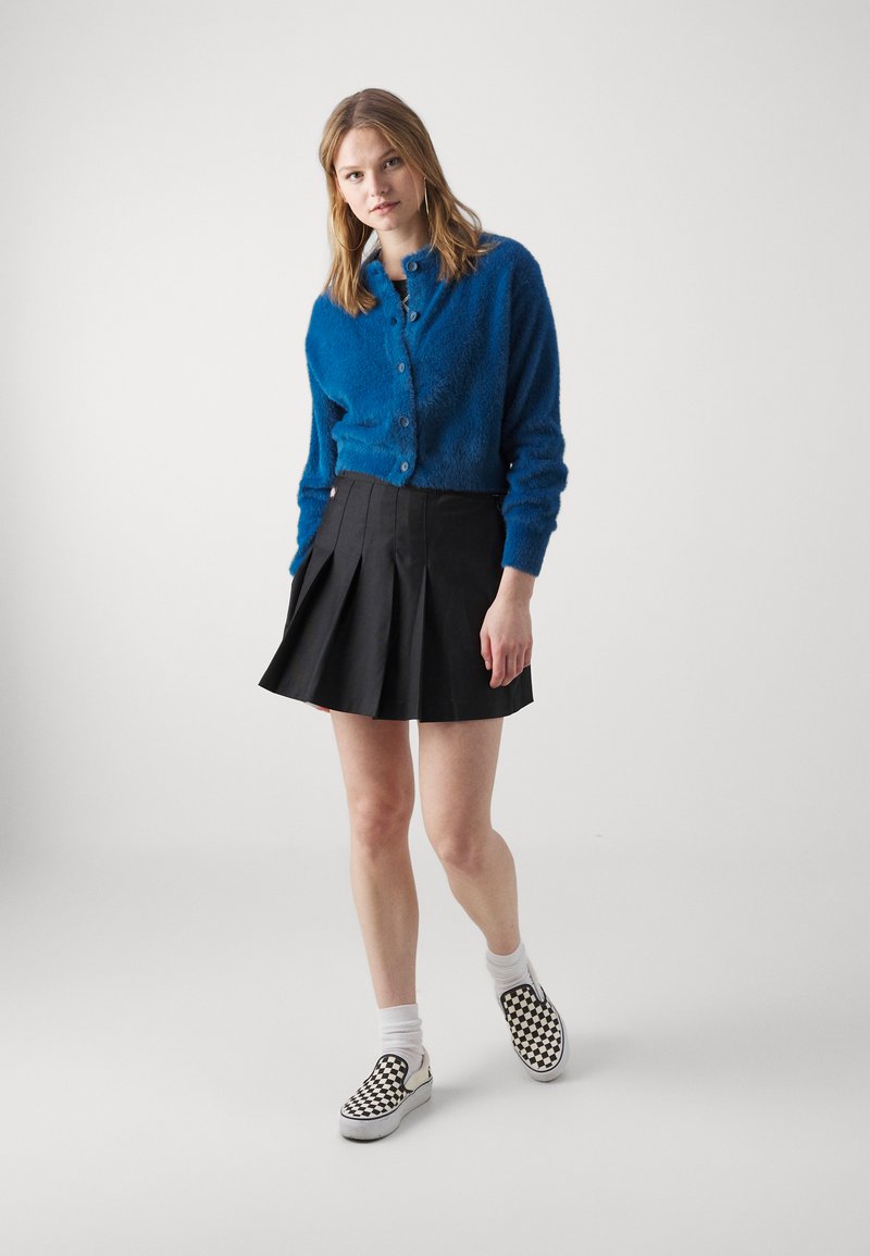 Cardigan blu fluffy, gonna nera plissettata, calze bianche e scarpe slip-on a scacchi bianchi e neri. Un outfit semplice e casual con colori distintivi.