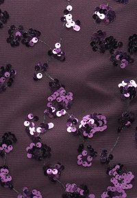 Μωβ ύφασμα από δίχτυ διακοσμημένο με μαύρες και λεβαντίνες παγιέτες τοποθετημένες σε ανθικά σχέδια, δημιουργώντας μια υφή με λάμψη.