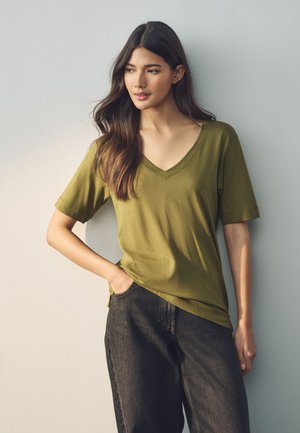 Kobieta z długimi ciemnymi włosami, ubrana w oliwkowo zielony T-shirt z dekoltem w serek i czarne dżinsy, stojąca z jedną ręką w kieszeni przy jasnej ścianie.