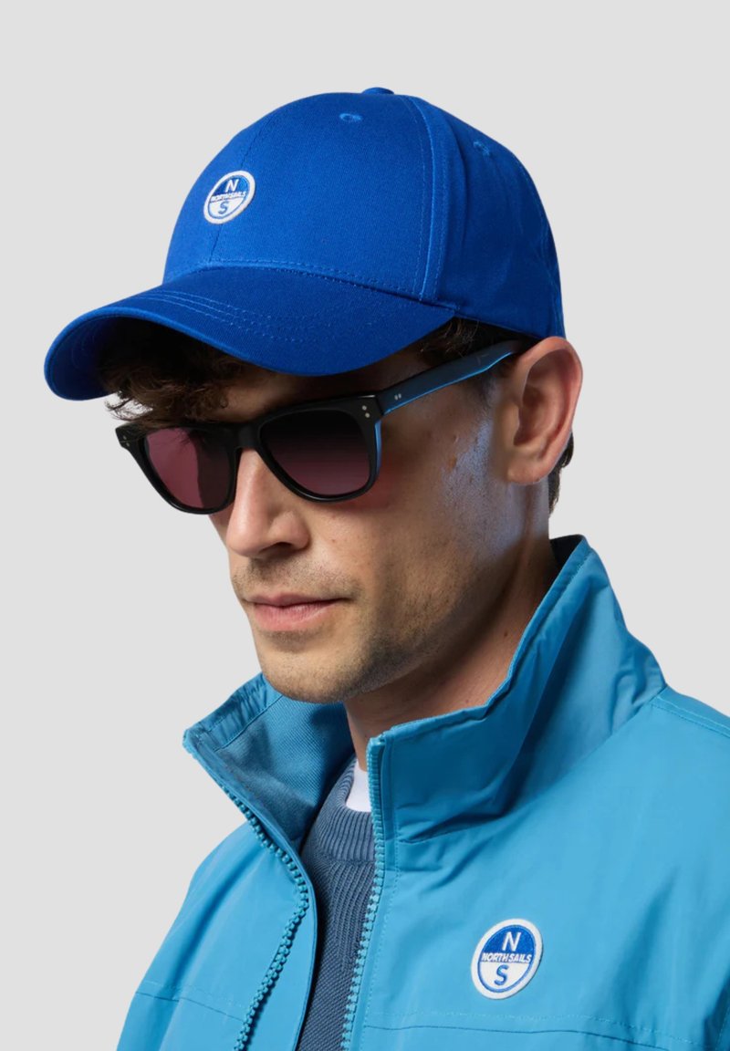Cappellino da baseball blu con un logo sulla parte anteriore, abbinato a occhiali da sole neri con lenti sfumate e a una giacca zip teal.