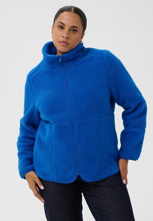Femme portant une veste polaire bleu vif zippée avec un col montant et un pantalon sombre sur un fond clair uni.