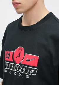 Μαύρο βαμβακερό μπλουζάκι με γραφικό σχέδιο σε κόκκινες αποχρώσε ις, που περιλαμβάνει τον αριθμό "23", το λογότυπο Jumpman και το κείμενο "Jordan Brand".