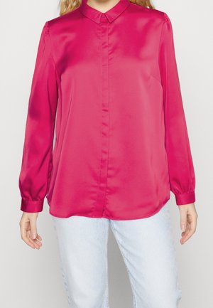 Vrouw draagt een felroze satijnen blouse met lange mouwen en knopen, gecombineerd met lichtblauwe spijkerbroek, gefotografeerd van nek tot heupen tegen een effen achtergrond.