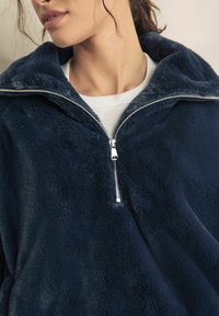 Pulover din fleece bleu marin cu guler înalt, fermoar pe jumătate în față și textură moale. Prezintă un design simplu, fără modele vizibile.