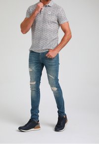 Kortärmad polo med ett grått och vitt triangelmönster, i kombination med slitna blåjeans och marinblå sneakers med bruna accenter.