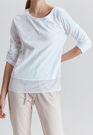 Femme portant un haut blanc à manches longues superposé avec un ourlet transparent et un pantalon beige à cordon, debout avec une main sur la hanche sur fond uni.