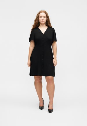 Fiatal nő hosszú göndör hajjal, fekete, gombos, térdig érő ruhában és fekete magas sarkú cipőben, fehér háttér előtt állva.