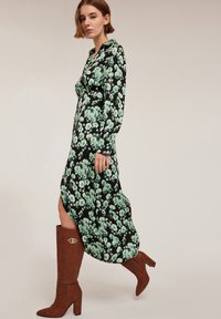 Rochie maxi florală neagră cu un model floral verde, având guler și mâneci lungi. Cizme maro până la genunchi cu un accent decorativ auriu.