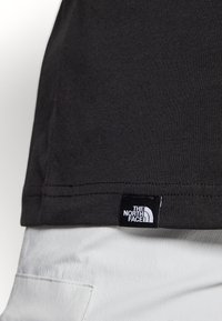 Czarna bawełniana koszulka z wyszywaną krawędzią i metką z białym logo "The North Face". Kontrastująca z jasnymi spodniami.