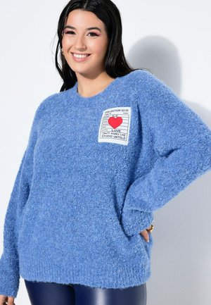 Smilende kvinde med langt mørkt hår iført en blå, fluffy sweater med en hvid lap, der har et rødt hjerte og tekst på brystet.