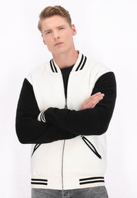 Jachetă bomber albă și neagră, cu fermoar în față, guler și manșete ribborate cu dungi, buzunare laterale și un corp solid negru.