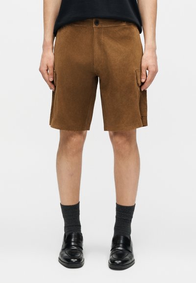Homme portant un short cargo en daim marron, des mocassins noirs et des chaussettes gris foncé, debout devant un fond clair uni.