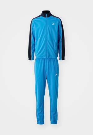 Jachetă cu fermoar și pantaloni elastici asortați, dintr-un material neted, de culoare albastră, cu accente negre. Logo-ul Nike este vizibil.