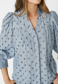 Blue and white striped blouse with a v-neck, featuring dark circular patterns. Puff sleeves and textured fabric add detail.