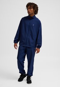 Navyblauwe trainingsoutfit van gladde stof, met een ritsjack met zakken en bijpassende broek. Zwarte sportschoenen maken de look compleet.