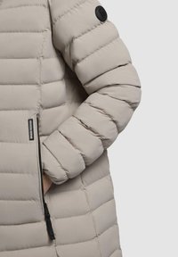 Giacca imbottita beige con tasca con zip e polsini a coste. Il tessuto è liscio con una finitura opaca e include una toppa circolare con logo.