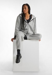 Felpa grigia con zip e pantaloni della tuta abbinati in una morbida texture. Il modello indossa una maglietta nera e sneaker nere. Seduto su un piedistallo bianco.