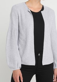 Cárdigan abierto de color gris claro con mangas abullonadas largas, usado sobre una camiseta negra y pantalones negros.