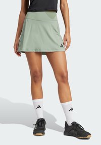 Falda atlética verde claro con cintura plana, hecha de un tejido suave. Presenta un logo de Adidas cerca del dobladillo. Combinada con zapatos negros y calcetines blancos.