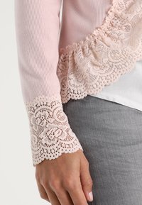 Ljusrosa stickad tröja med en volangkant i spets längs nederkanten och ärmarna, prydd med intrikata blommiga spetsmönster vid manschetterna.