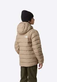 Geacă matlasată bej cu design matlasat orizontal, glugă atașată și logo alb în partea superioară stângă. Asortată cu pantaloni verzi și o căciulă neagră.