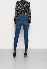 Pantalones vaqueros ajustados de cintura alta en azul con una textura suave, cinco bolsillos y un diseño clásico, combinados con una blusa negra estampada y zapatos de tacón.
