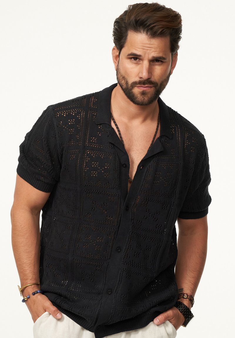 Schwarzes, gestricktes Kurzarmshirt mit einem strukturierten, offenen Muster. Verfügt über eine knopflose Vorderseite und einen Kragen. Akzentuiert mit schwarzen Knöpfen.