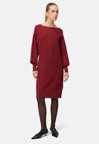 Burgunderfarbenes Strickkleid mit weitem Ausschnitt, langen Ärmeln, strukturiertem Stoff und gerader Silhouette, kombiniert mit transparenten schwarzen Strumpfhosen und spitzen Ballerinas.