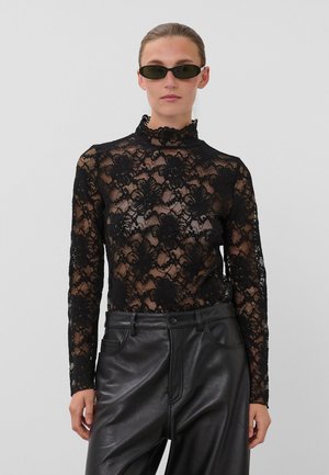Femme portant un haut à manches longues en dentelle florale noire, un pantalon en cuir noir et de petites lunettes de soleil noires, les cheveux tirés en arrière sur un fond clair uni.