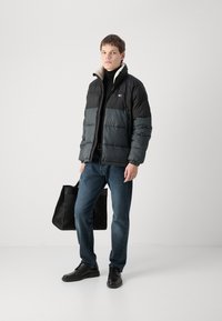 Giubbotto imbottito con blocchi di colore nero e grigio, polsini elasticizzati e zip sul davanti, abbinato a jeans scuri e scarpe nere, mentre tiene una borsa nera con texture.