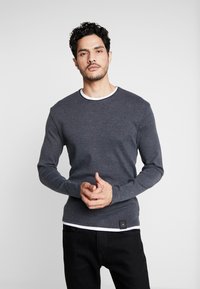 Graues Langarmshirt mit weißem Rundhalsausschnitt, aus einem strukturierten Stoff gefertigt, mit einem kleinen Logo-Patch am Saum.