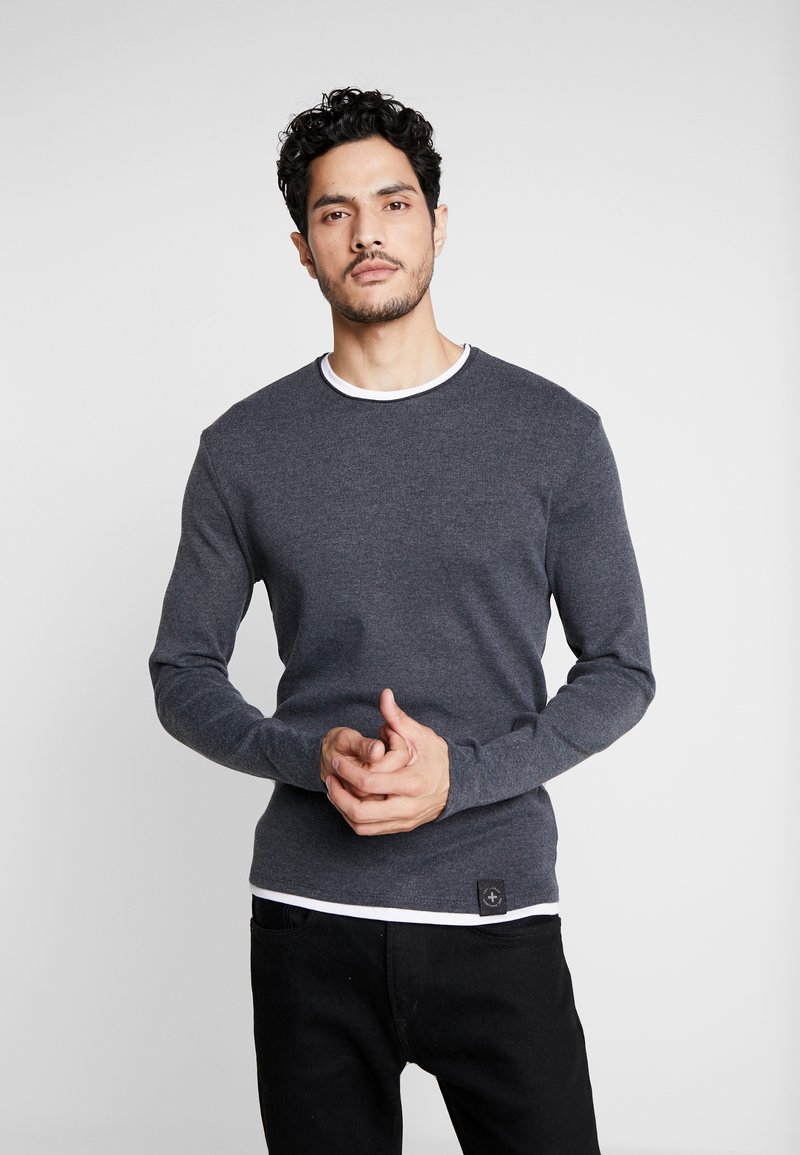 Graues Langarmshirt mit weißem Rundhalsausschnitt, aus einem strukturierten Stoff gefertigt, mit einem kleinen Logo-Patch am Saum.