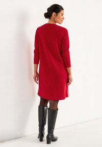 Rode ribgebreide jurk met driekwart mouwen, een ronde halslijn en een flatterende fit. Combinatie met zwarte leren laarzen tot de knie.