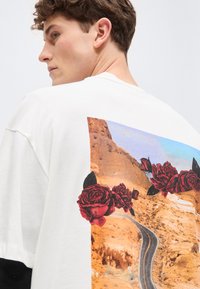 Νεαρός άνδρας με κοντά, σγουρά μαλλιά, που φοράει ένα λευκό πουκάμισο με γραφικό στοιχείο στον πίσω μέρος που απεικονίζει δρόμο στην έρημο και ένα κόκκινο τριαντάφυλλο.