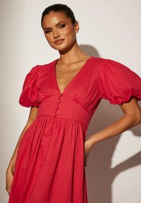 Röd klänning med puffärmar och djup V-ringning. Texturerad tyg med ett vertikalt randmönster. Har en rad knappar fram.