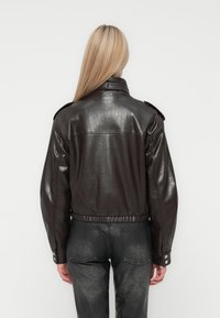 Kvinde med langt blond hår iført en mørkebrun læderjakke og slidte sorte jeans, vist fra ryggen mod en ensfarvet baggrund.