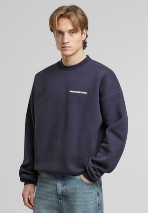 Suedou oversized de culoare bleumarin, cu guler raglan, având un logo roz „PROHIBIT” pe piept și un material texturat.