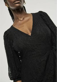 Black lace dress featuring a deep V-neck, three-quarter puff sleeves, and a tie waist. The fabric has a floral pattern and a textured finish.