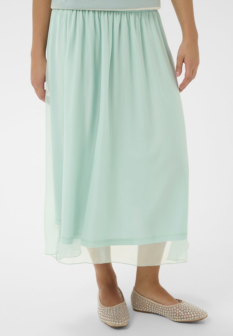 Jupe en mousseline vert menthe avec une taille élastique, présentant un design fluide et une texture superposée. Associée à des ballerines beige ornées.