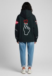 Felpa nera con una grafica di una mano e un cuore sulla schiena. Presenta dettagli rossi e bianchi, maniche lunghe e una tasca frontale. Indossata con pantaloni blu chiaro.