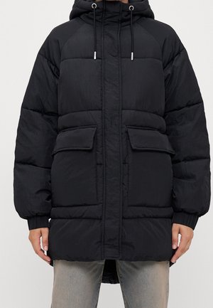 Personne portant une parka noire matelassée avec capuche, cordons de serrage, deux grandes poches à rabat à l'avant et des poignets élastiques, sur un jean de couleur claire.