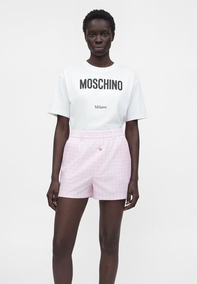 Femme portant un t-shirt blanc Moschino Milano et un short à carreaux rose clair taille haute, debout devant un fond blanc uni.