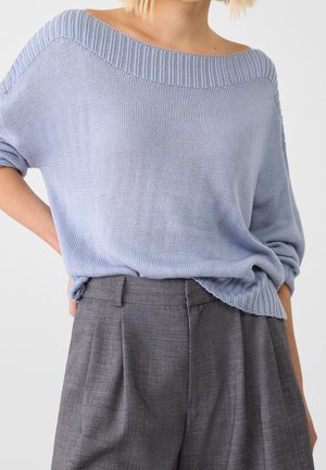 Kobieta ubrana w jasnoniebieski, odkrywający ramiona sweter z dzianiny włożony do wysokich, szarych spodni z plisami, z rękami spoczywającymi z tyłu.