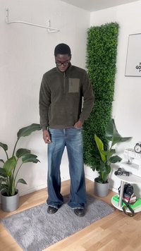 Mand iført briller, olivengrøn fleecepullover, blå vide jeans og sorte sko, der står på et gråt tæppe i et rum med planter og kameratur.